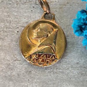 May include: A gold-colored pendant with a circular design featuring a profile and text. The pendant has a loop at the top for a chain. The background is a textured gray surface.