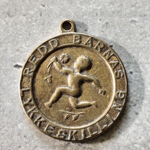 May include: A vintage bronze pendant with the text "REDD BARNAS LYKKESKILLING" encircling a central image of a running child holding a flower. The pendant has a loop at the top for a chain.