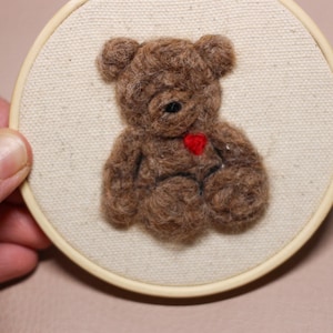 May include: A brown felt teddy bear with a red heart on its chest, framed in a white embroidery hoop.