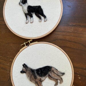 May include: Two circular wooden embroidery hoops containing needle-felted dog portraits on off-white fabric. The top hoop features a black and white dog standing in profile. The bottom hoop displays a black and tan dog standing in profile.