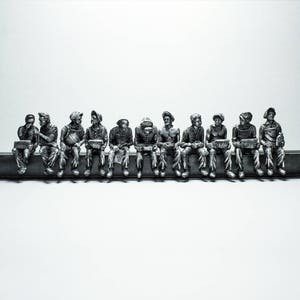 Può includere: Scultura monocromatica di undici figure sedute su una trave d'acciaio. Ogni figura, vestita con abiti da lavoro, tiene una scatola per il pranzo. La scultura è ambientata su uno sfondo bianco.
