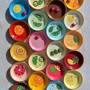May include: A collection of small, colorful ceramic bowls, each adorned with a different fruit or garnish design. The bowls feature vibrant colors like blue, yellow, red, and pink, with detailed illustrations of lemons, cherries, and olives.