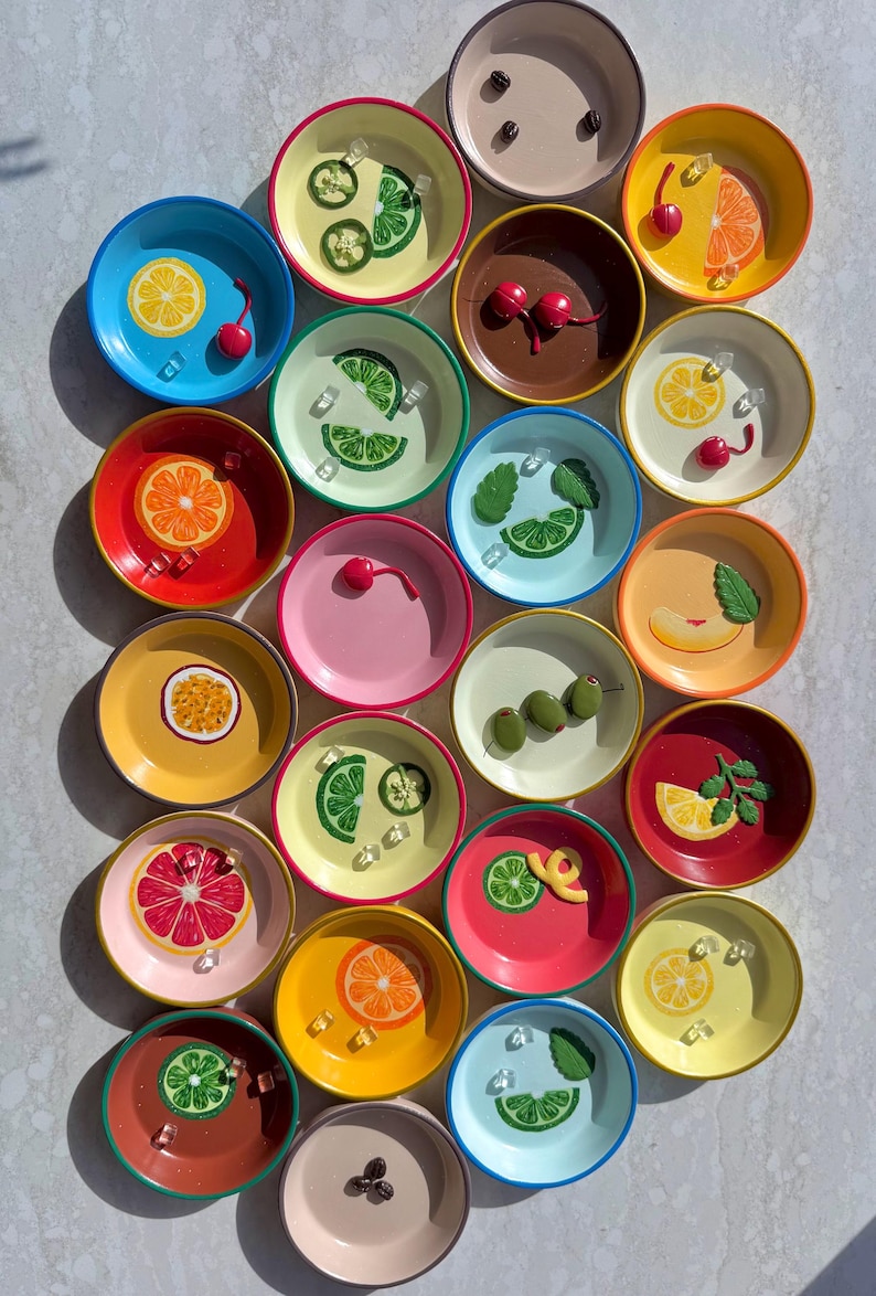 May include: A collection of small, colorful ceramic bowls, each adorned with a different fruit or garnish design. The bowls feature vibrant colors like blue, yellow, red, and pink, with detailed illustrations of lemons, cherries, and olives.