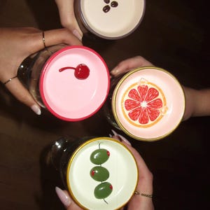 May include: Overhead shot of four cocktail glasses, each with a unique garnish. One glass has three coffee beans, another a cherry, a third a grapefruit slice, and the last, three olives. The glasses are held by hands.