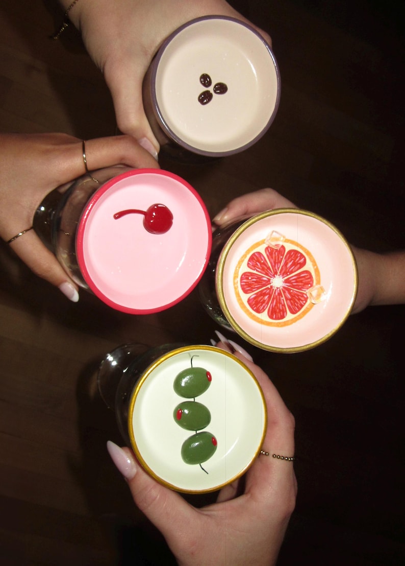 May include: Overhead shot of four cocktail glasses, each with a unique garnish. One glass has three coffee beans, another a cherry, a third a grapefruit slice, and the last, three olives. The glasses are held by hands.
