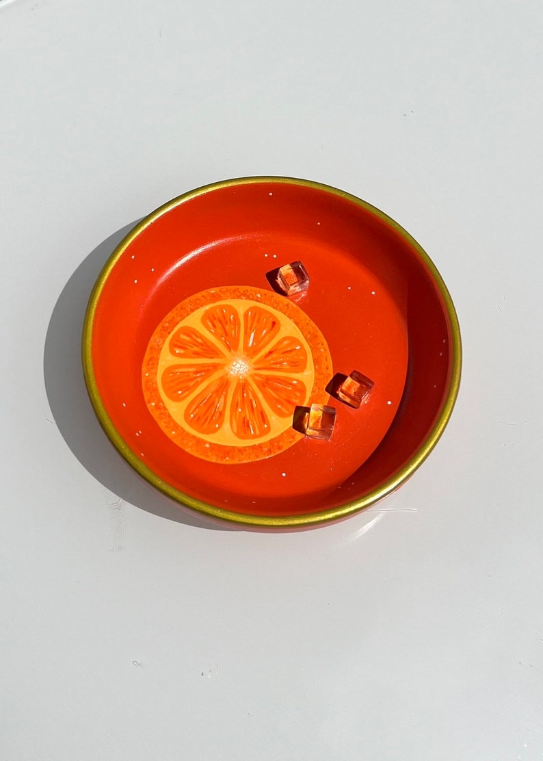 May include: A round, orange-colored dish with a gold rim and a painted orange slice design. The dish has three clear, square-shaped decorative pieces. The dish is on a white surface.
