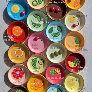 May include: A collection of colorful, small ceramic plates, each decorated with a different cocktail design. The plates feature various fruits, garnishes, and cocktail names like "Blue Lagoon" and "Tequila Sunrise".