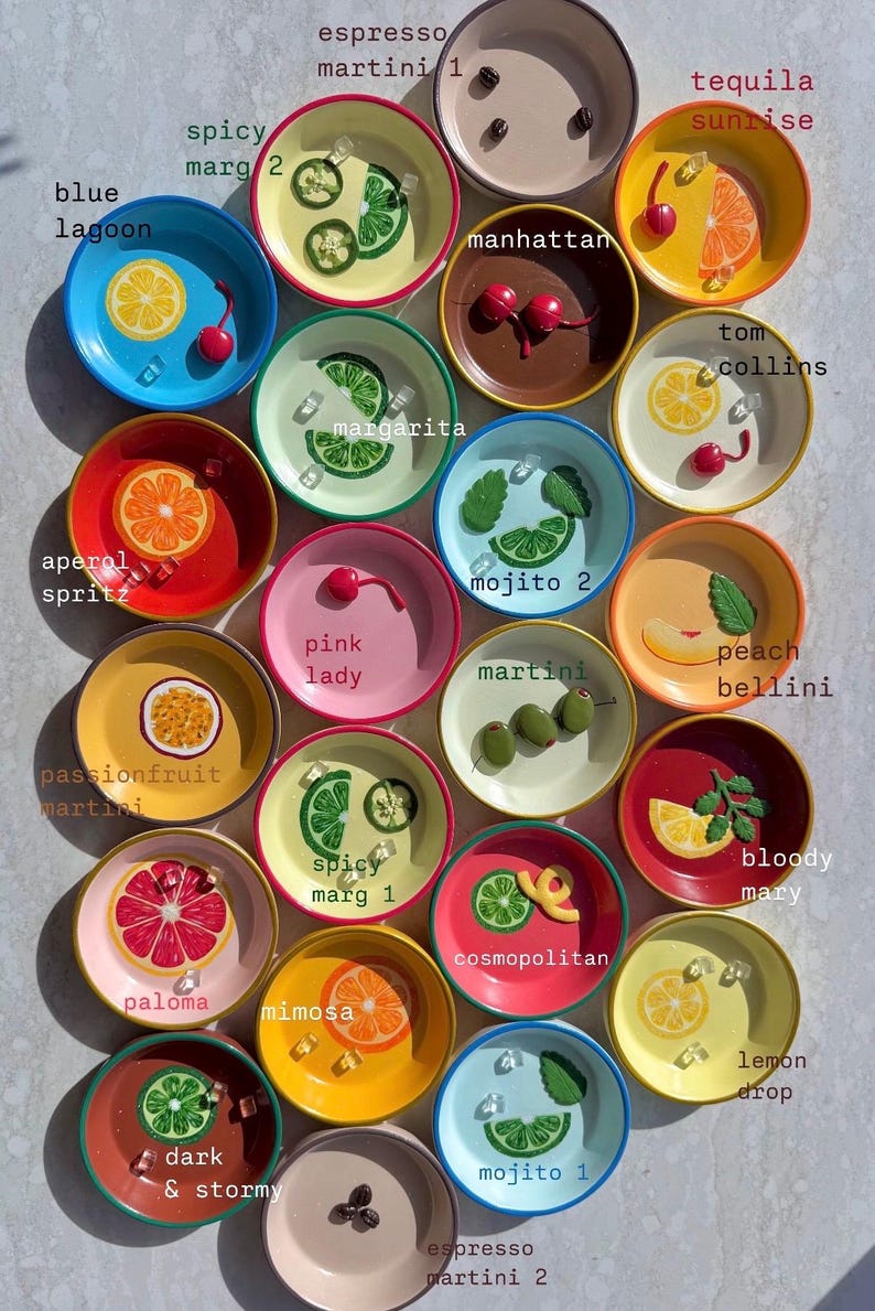 May include: A collection of colorful, small ceramic plates, each decorated with a different cocktail design. The plates feature various fruits, garnishes, and cocktail names like "Blue Lagoon" and "Tequila Sunrise".