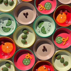 May include: Overhead shot of colorful small bowls with various garnishes. The bowls are in shades of red, pink, green, and tan, each with a different design, including orange slices, lime wedges, olives, and coffee beans. Some bowls have ice cubes and orange zest.