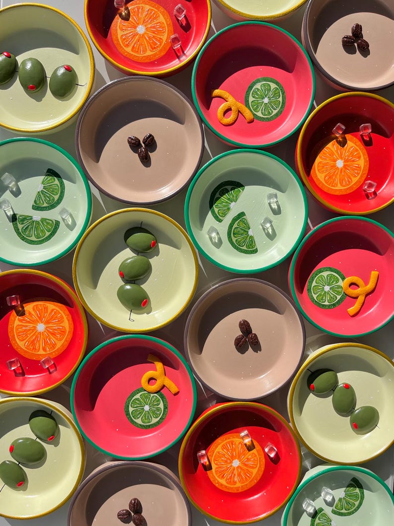 May include: Overhead shot of colorful small bowls with various garnishes. The bowls are in shades of red, pink, green, and tan, each with a different design, including orange slices, lime wedges, olives, and coffee beans. Some bowls have ice cubes and orange zest.