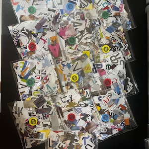 May include: Nine clear plastic bags filled with colorful paper scraps. The scraps are cut into various shapes and sizes, and some have letters printed on them. The bags are arranged in a pyramid shape on a black surface.