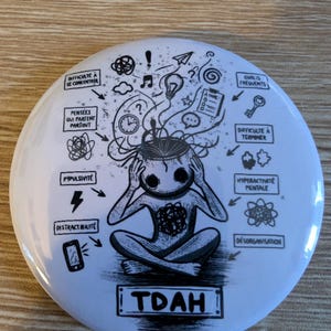 Peut inclure: Bouton blanc avec une illustration en noir et blanc d'une personne avec des bulles de pensée et le texte "TDAH". L'illustration comprend des mots comme "Impulsivité" et "Difficulté à se concentrer".