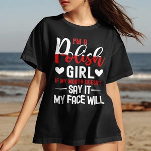 May include: Black t-shirt with the text "I'M A Polish GIRL" in white and red lettering. The design includes two white hearts and the phrase "IF MY MOUTH DOESN'T SAY IT - MY FACE WILL". The shirt is worn by a person on a beach.