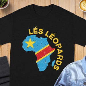 May include: A black short-sleeve t-shirt featuring a distressed graphic of the African continent filled with the Democratic Republic of the Congo flag colors, a yellow star, and the yellow text LÉS LÉOPARDS arched over the top right side.