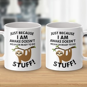 May include: Two white ceramic mugs with a humorous sloth design. Each mug features the text "JUST BECAUSE I AM AWAKE DOESN'T MEAN I'M READY TO DO STUFF!" with a cartoon sloth hanging from a branch.