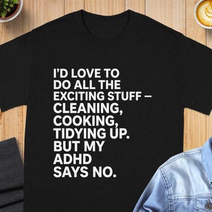 Op de afbeelding: Een zwart t-shirt met korte mouwen en witte tekst: I'D LOVE TO DO ALL THE EXCITING STUFF - CLEANING, COOKING, TIDYING UP. BUT MY ADHD SAYS NO. Het shirt heeft een ronde hals en een standaard pasvorm, gepresenteerd op een plat oppervlak.