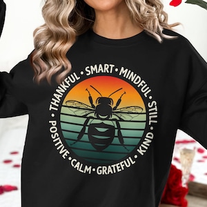 May include: Black sweatshirt featuring a circular design with a bee illustration. The design includes the words "Thankful, Smart, Mindful, Still, Kind, Grateful, Calm, Positive" in white. The bee is set against a sunset-colored background.