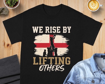 May include: A black short-sleeve t-shirt featuring a graphic of a rugby lineout silhouette over the flag of England. The text WE RISE BY LIFTING OTHERS is printed in tan and white lettering above and below the central flag and player graphic.