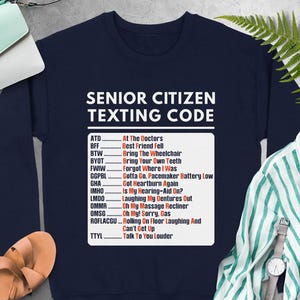 May include: Navy blue sweatshirt with white text: "SENIOR CITIZEN TEXTING CODE." The text includes common abbreviations and their meanings, such as "ATD - At The Doctors" and "BTW - Bring The Wheelchair." The sweatshirt is surrounded by other items.