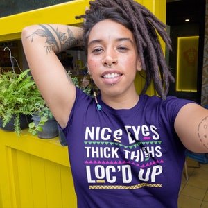 May include: Purple t-shirt with the text "Nice Eyes Thick Thighs Loc'd Up" in colorful block letters. The shirt has a geometric pattern around the text.