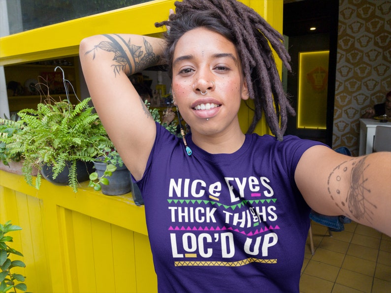 May include: Purple t-shirt with the text "Nice Eyes Thick Thighs Loc'd Up" in colorful block letters. The shirt has a geometric pattern around the text.