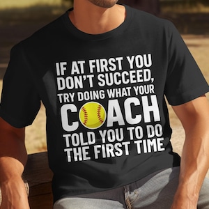 May include: Black t-shirt with white text that reads "IF AT FIRST YOU DON'T SUCCEED, TRY DOING WHAT YOUR COACH TOLD YOU TO DO THE FIRST TIME." A yellow softball graphic is in the word "COACH".