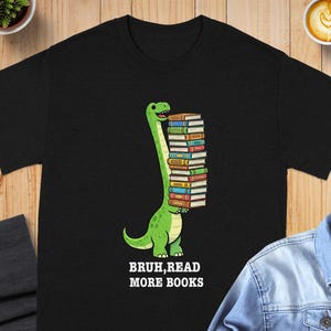 May include: Black t-shirt featuring a green dinosaur carrying a stack of colorful books. The dinosaur has a long neck and a friendly expression. The text "BRUH, READ MORE BOOKS" is printed below the dinosaur.
