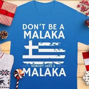 May include: A blue t-shirt with the text "DON'T BE A MALAKA" above a Greek flag design and the text "NOBODY LIKES A MALAKA" below. The shirt is surrounded by holiday decorations, including a gift box, ornaments, and a sweater.