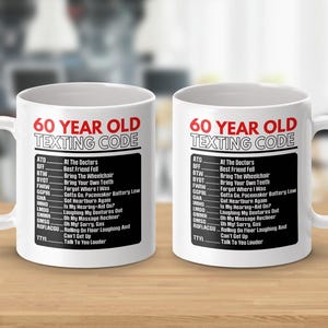 May include: Two white ceramic mugs with the text "60 YEAR OLD TEXTING CODE" in red and black. The mugs have a list of texting abbreviations and their meanings, such as "BTW - Bring The Wheelchair."