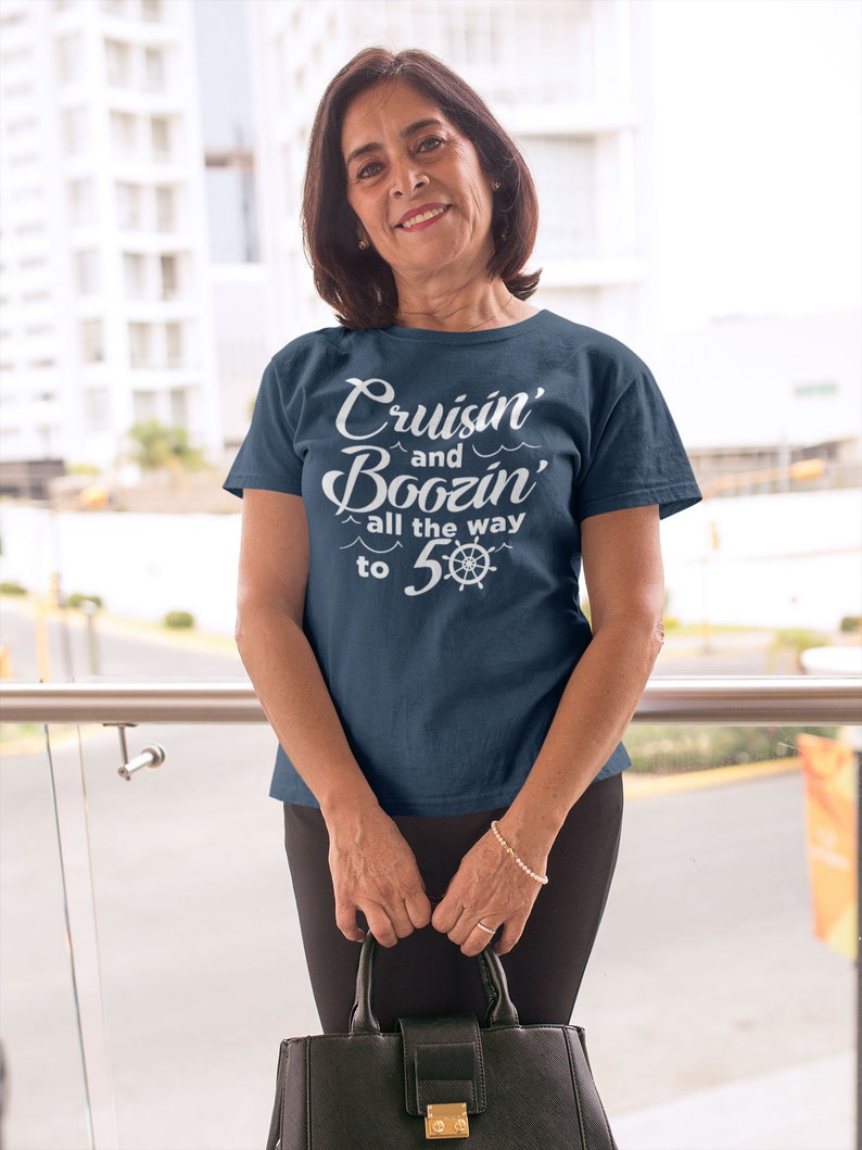 K&ouml;nnte beinhalten: Eine Frau tr&auml;gt ein marineblaues T-Shirt mit dem Text "Cruisin' and Boozin' all the way to 5-0" in Wei&szlig;. Das Shirt zeigt ein nautisches Rad-Motiv.