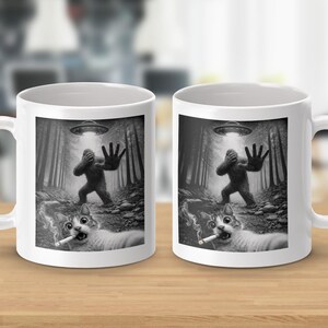 May include: Two white ceramic mugs with a humorous black and white graphic. The design features a Bigfoot-like creature, a UFO, and a cat smoking a cigarette in a forest setting. The mugs have a standard handle and a glossy finish.