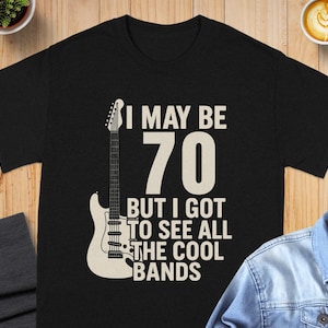 May include: Black t-shirt with the text "I MAY BE 70 BUT I GOT TO SEE ALL THE COOL BANDS" and a white guitar graphic. The shirt is on a wooden surface with a denim jacket, a cup of coffee, and a small plant.
