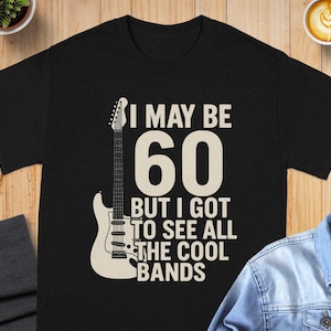 May include: Black t-shirt with the text "I MAY BE 60 BUT I GOT TO SEE ALL THE COOL BANDS" and a white guitar graphic. The shirt is displayed on a wooden surface.