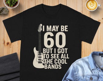 May include: Black t-shirt with the text "I MAY BE 60 BUT I GOT TO SEE ALL THE COOL BANDS" and a white guitar graphic. The shirt is displayed on a wooden surface.