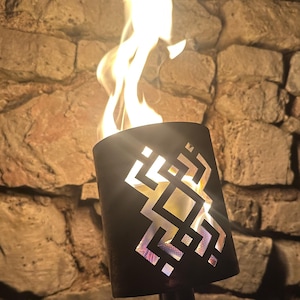 May include: A black metal torch with a geometric cut-out design, emitting bright flames. The torch is mounted on a black pole, set against a backdrop of a stone wall. The flames are yellow and orange.