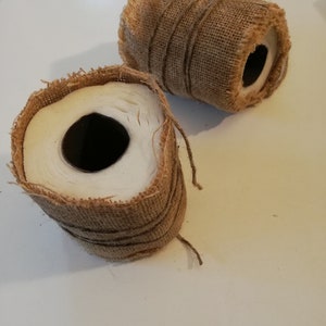 Pode incluir: Dois rolos de papel higiênico embrulhados em tecido de juta marrom. Os rolos estão sobre uma superfície branca.