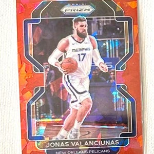 May include: A basketball trading card featuring Jonas Valanciunas of the New Orleans Pelicans. The card has a red and blue design with the Panini Prizm logo. Valanciunas is shown in his Memphis Grizzlies uniform, holding a basketball.