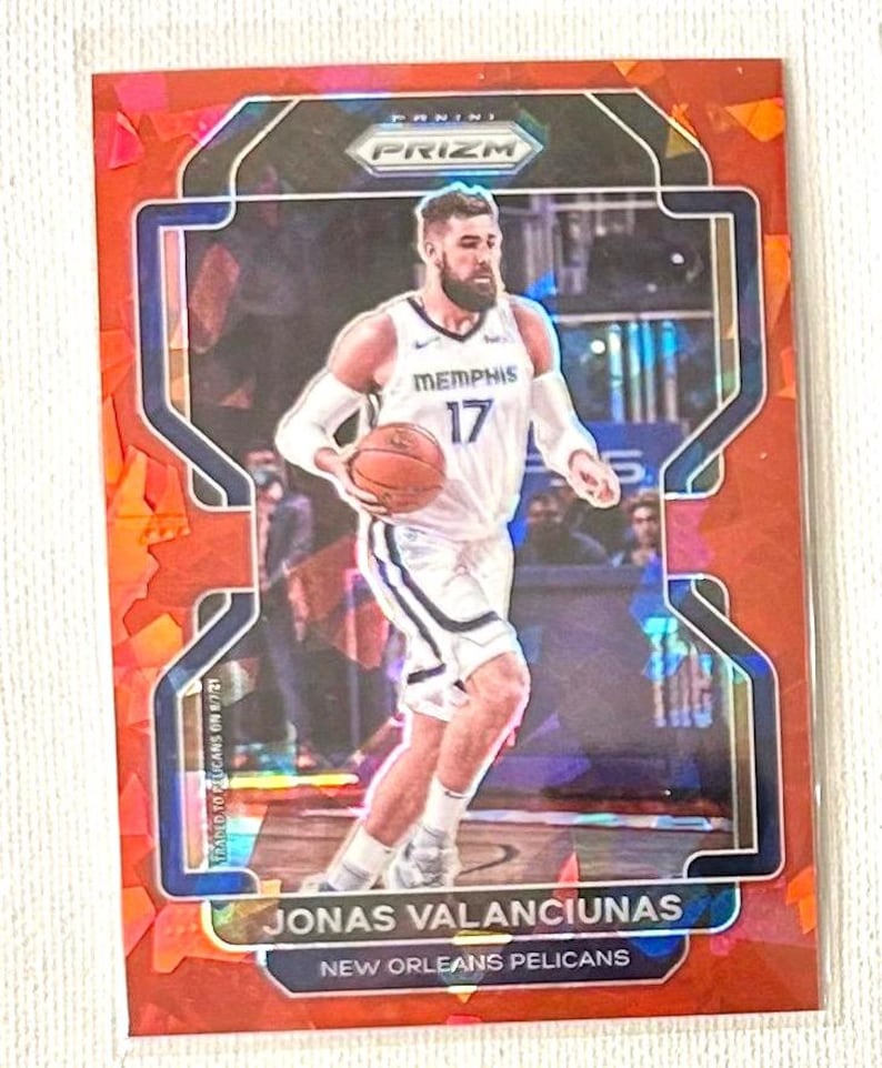 May include: A basketball trading card featuring Jonas Valanciunas of the New Orleans Pelicans. The card has a red and blue design with the Panini Prizm logo. Valanciunas is shown in his Memphis Grizzlies uniform, holding a basketball.