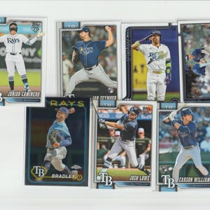 Può includere: Una collezione di sette figurine di baseball dei Tampa Bay Rays con i giocatori Junior Caminero, Ian Seymour e altri in pose d'azione. Ogni carta mostra il logo della squadra, il nome del giocatore e il marchio del 75° anniversario di Topps.