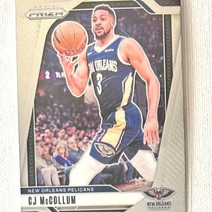 May include: A basketball trading card featuring CJ McCollum of the New Orleans Pelicans. The card has a silver border and includes the team logo and player's name. McCollum is shown in his team uniform, holding a basketball.