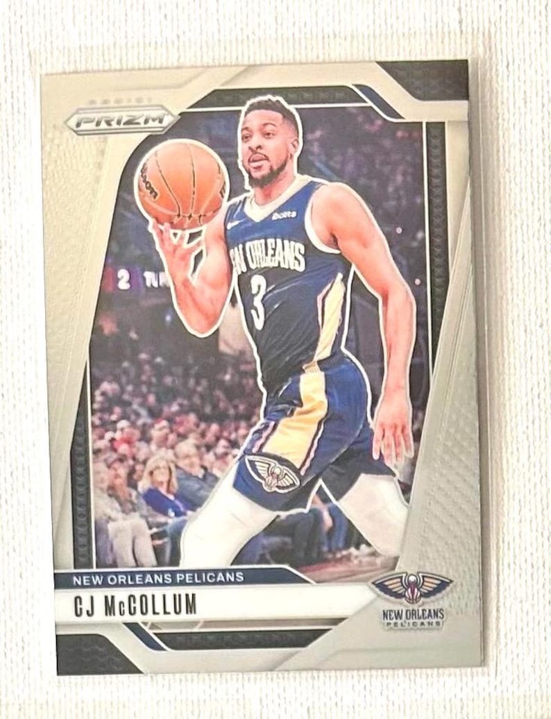 May include: A basketball trading card featuring CJ McCollum of the New Orleans Pelicans. The card has a silver border and includes the team logo and player's name. McCollum is shown in his team uniform, holding a basketball.