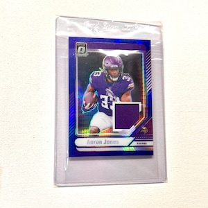 May include: A trading card featuring Aaron Jones in a purple and white uniform, with the text "Aaron Jones" and "Vikings". The card is encased in a clear plastic sleeve.
