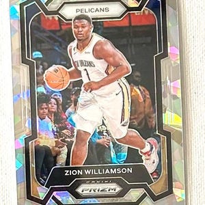 May include: A basketball trading card featuring Zion Williamson of the New Orleans Pelicans. The card has a holographic background and a black and silver frame. The player is wearing a white jersey and holding a basketball.