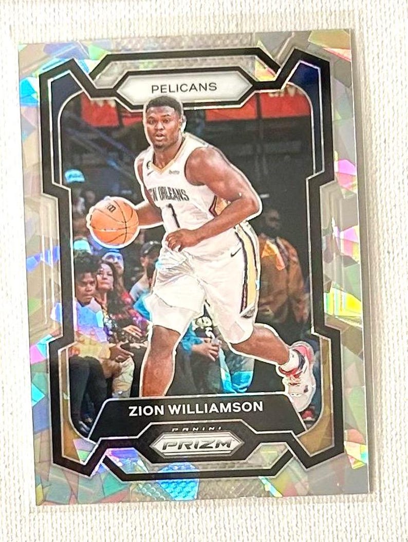 May include: A basketball trading card featuring Zion Williamson of the New Orleans Pelicans. The card has a holographic background and a black and silver frame. The player is wearing a white jersey and holding a basketball.