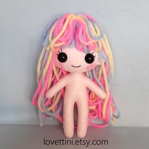 Puede incluir: Una mu&ntilde;eca de fieltro hecha a mano con el pelo largo de hilo de colores. La mu&ntilde;eca tiene un cuerpo rosa, ojos negros y una boca sonriente.