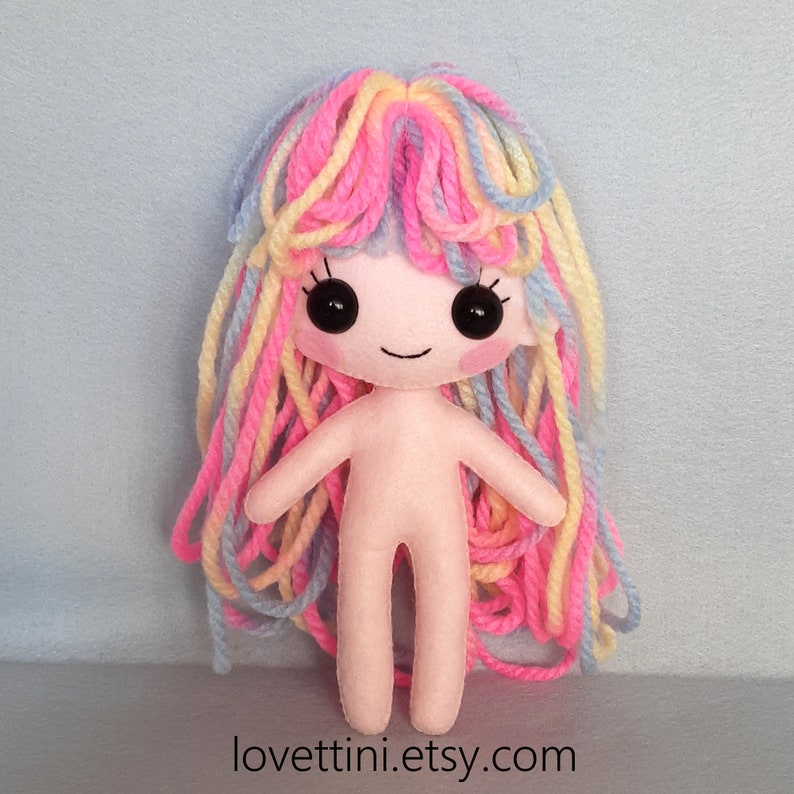 Puede incluir: Una mu&ntilde;eca de fieltro hecha a mano con el pelo largo de hilo de colores. La mu&ntilde;eca tiene un cuerpo rosa, ojos negros y una boca sonriente.
