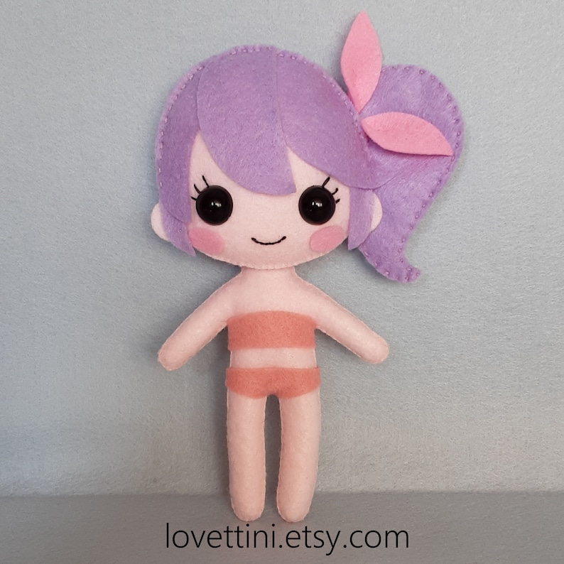 Puede incluir: Una mu&ntilde;eca de fieltro con cabello morado, un lazo rosa y un traje de ba&ntilde;o color melocot&oacute;n. La mu&ntilde;eca tiene una cara sonriente con ojos negros y mejillas rosadas.