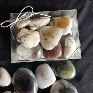 May include: A collection of smooth, polished stones in various shades of gray, white, and brown, displayed in a clear plastic box with a silver ribbon.