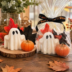 May include: Two white ghost-shaped candles with black eyes, each paired with a small orange glittery pumpkin and red and black fabric maple leaves. One set is unwrapped, while the other is inside a clear plastic bag tied with a black ribbon.