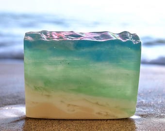 May include: A rectangular bar of soap with a layered design resembling a beach scene. The soap features a sandy base, transitioning to green and blue hues, and a textured top, evoking ocean waves. The soap is sitting on a sandy surface.